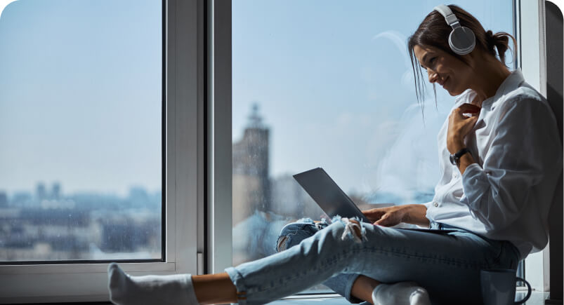 Young woman wearing headphones sitting by a window, using a laptop with a smile