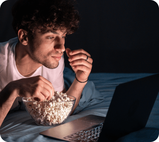 Young man with curly hair watching a movie on his laptop while eating popcorn in a dimly lit room