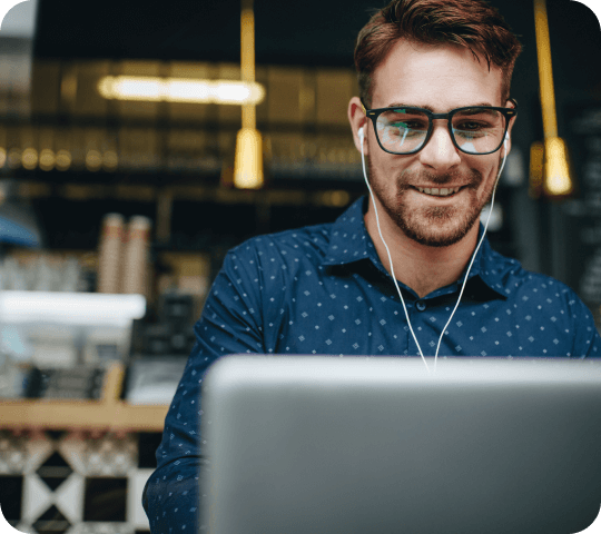 Smiling man with glasses and earbuds using a laptop in a café