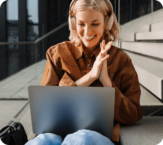 Smiling blonde woman with headphones using a laptop while sitting on outdoor steps