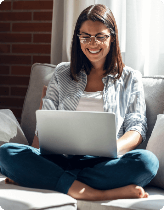 Smiling woman with glasses sitting cross-legged on a couch while using a laptop